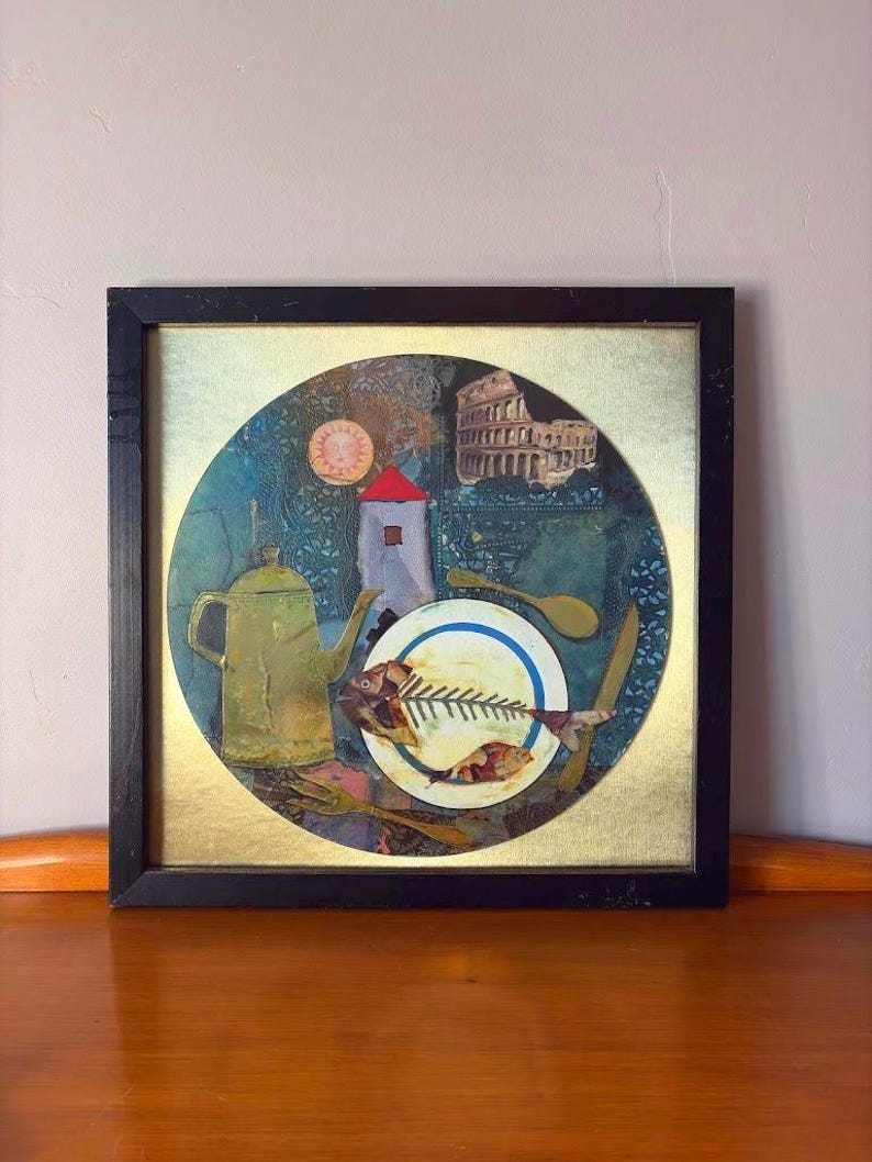 Framed mixed media collage artwork featuring a central white plate with a fish skeleton, surrounded by a golden teapot, wooden cutlery, and background motifs including a red-roofed tower, the Roman Colosseum, and a stylized sun. The frame is black with signs of wear, placed on a wooden surface against a light wall.