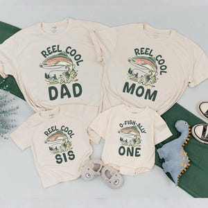 May include: Cream-colored family matching t-shirts and a onesie with a fishing theme. The shirts have a graphic of a fish with the words "REEL COOL DAD", "REEL COOL MOM", and "REEL COOL SIS". The onesie says "O-FISH-ALLY ONE".