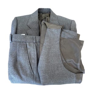 Könnte beinhalten: Ein grauer Wollanzug, bestehend aus Blazer, Weste und Hose, ist ordentlich angeordnet. Der Blazer hat ein fallendes Revers und ein Einstecktuch. Weste und Hose sind sichtbar. Der Anzug ist ein klassischer Stil.
