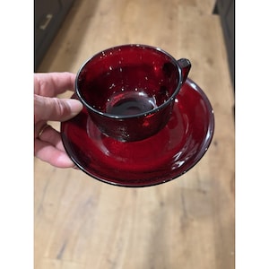 May include: A deep red glass teacup and saucer. The cup has a small handle and sits on a matching saucer. The set is made of textured glass. The set is held in a person's hand.