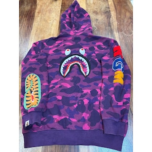 May include: A purple and pink camouflage print hoodie featuring a large shark mouth graphic on the back. The right sleeve displays a tiger graphic, while the left sleeve features the text WGM in large, colorful, embroidered block letters.