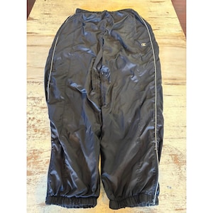 May include: Black nylon track trousers featuring an elasticated waistband and a white Champion logo on the upper right thigh. The trousers include thin white piping running down the outer legs and zips at the bottom of each leg cuff for adjustable fit.