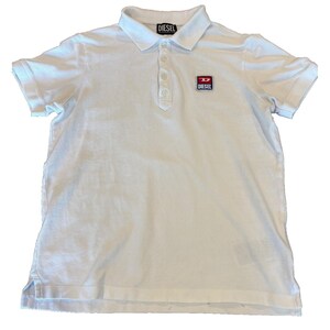 Könnte beinhalten: Ein weißes kurzärmeliges Poloshirt mit klassischem Kragen, einer Knopfleiste mit vier Knöpfen und einem kleinen rechteckigen Aufnäher auf der linken Brust, der das rot-weiß-blaue Diesel-Logo zeigt. Das Innenetikett am Kragen trägt ebenfalls den Markennamen Diesel.