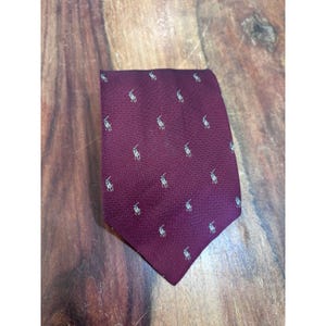 Peut inclure: Une cravate bordeaux foncé ornée d'un motif répétitif de petits logos de joueur de polo brodés en blanc. Le tissu présente une texture tissée en diagonale subtile sur toute la surface de la cravate, qui est pliée en forme pointue.