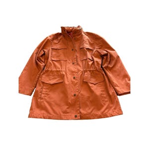 Könnte beinhalten: Eine orangefarbene Jacke mit hohem Kragen und mehreren Taschen. Die Jacke hat eine Taille mit Kordelzug und Knopfverschlüssen vorne. Die Ärmel sind lang und die Jacke scheint aus leichtem Material zu sein. Größe Medium.