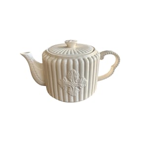 Könnte beinhalten: Cremefarbene Keramik-Teekanne mit Deckel und Henkel. Die Teekanne hat vertikale Rippendetails und ein dekoratives Blumenemblem auf der Vorderseite. Der Griff und der Ausguss sind gebogen, und der Deckel hat einen kleinen Knopf.
