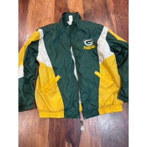 Może przedstawiać: Wiatrówka Green Bay Packers z wzorem typu color-block w kolorach ciemnej zieleni, jaskrawej żółci i bieli. Kurtka posiada zamek błyskawiczny na całej długości, stójkę, długie rękawy z elastycznymi mankietami oraz logo Packers na piersi.