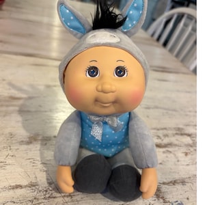 Peut inclure: Une poupée en peluche au visage rond, aux joues roses et aux yeux marron, portant une capuche grise sur le thème de l'âne avec des touffes de cheveux noirs et des oreilles à pois bleus. Le corsage est bleu à pois avec un ruban argenté.