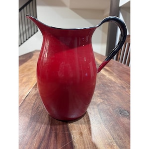 May include: A bright red enamel pitcher with a glossy finish, featuring a curved pouring spout and a solid black handle. The pitcher has a rounded body and a smooth, reflective surface, with a black rim accentuating the top edge of the vessel.
