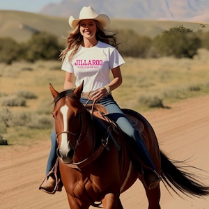 Peut inclure: Une femme portant un t-shirt blanc avec le texte "JILLAROO" et "BEING IS IN THE WAY" en lettres roses, chevauchant un cheval brun avec une bande blanche sur le visage. La femme porte un chapeau de cow-boy et un jean bleu.