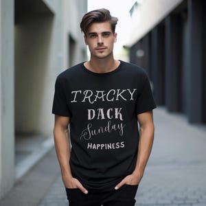 May include: Black crew neck t-shirt with the words "TRACKY DACK Sunday HAPPINESS" printed in white. The shirt is worn by a person standing outdoors with hands in pockets.