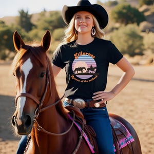 Peut inclure: Un t-shirt noir avec un graphique de coucher de soleil et le texte "Jillaroo's Queens of the outback". Une femme porte le t-shirt en chevauchant un cheval brun.