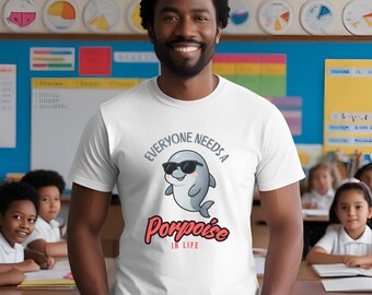 May include: White t-shirt featuring a cartoon dolphin wearing sunglasses. The shirt displays the text "Everyone Needs a Porpoise in Life". The image shows a person in a classroom environment.