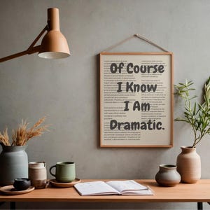 Puede incluir: Un cuadro enmarcado con el texto "Of Course I Know I Am Dramatic" cuelga de una pared gris. Un escritorio de madera exhibe una taza, jarrones y un cuaderno abierto. También se ven una lámpara de madera y vegetación.