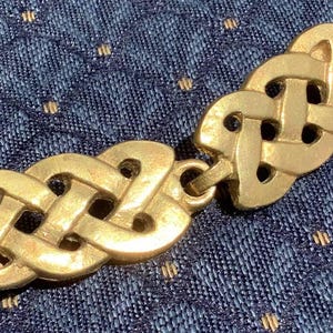 May include: Gold-colored Celtic knot clasp. The clasp features an intricate interwoven design, with two distinct sections connected by a small loop. The background is a textured blue fabric with small gold-colored dots.