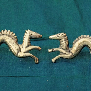Peut inclure: Deux figurines antiques en laiton représentant des hippocampes avec un corps serpentin et une tête de cheval. Les hippocampes se font face, avec un design texturé et détaillé. Le fond est un tissu de couleur sarcelle.