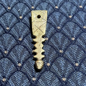 Puede incluir: Un colgante de metal dorado con un diseño geométrico. El colgante tiene una parte superior puntiaguda y una parte inferior redondeada. Está decorado con una serie de líneas y un pequeño agujero en el centro.