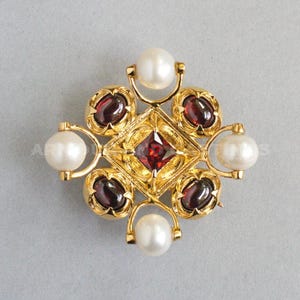 May include: A gold brooch featuring a central red gemstone in a diamond shape, encircled by six red gemstones and four white pearls. The brooch has an intricate, symmetrical design.