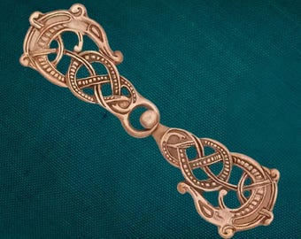 Puede incluir: Un broche para capa de metal en tono cobre con un intrincado diseño de nudos simétricos. El cierre consta de dos secciones alargadas y espejadas conectadas por un eslabón circular central con patrones entrelazados.