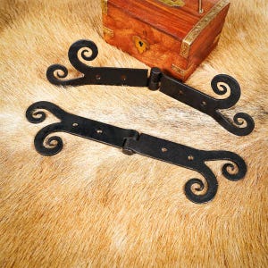 May include: Two black wrought iron hinges with decorative scrollwork ends are displayed on a textured, tan surface. A wooden chest with brass accents sits in the background. The hinges are designed for a rustic or vintage aesthetic.