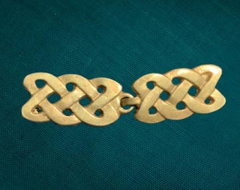 Puede incluir: Un broche de metal dorado para capa que presenta dos piezas simétricas con nudos entrelazados, conectadas por una bisagra central. El diseño consiste en bucles continuos y tejidos que forman un patrón repetitivo con espacios abiertos.