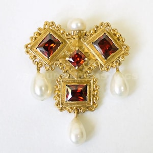 Peut inclure: Broche dorée ornée de pierres rouges carrées et losangiques, avec des perles en forme de goutte pendantes. La broche a une esthétique vintage, avec une petite perle au sommet. Le design est complexe et détaillé.