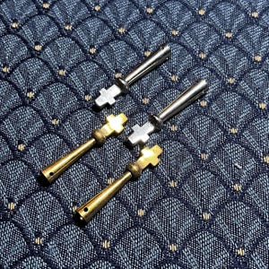 Puede incluir: Primer plano de cuatro colgantes metálicos en forma de cruz. Dos son de color plateado y dos dorados. Los colgantes tienen un diseño de cruz en la parte superior y un vástago cilíndrico. Los colgantes están sobre una tela azul oscuro estampada.