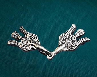 Puede incluir: Un broche de metal plateado para capa que consta de dos piezas simétricas con intrincados patrones de nudos en relieve. Cada pieza tiene un diseño de tres puntas en forma de ala, unidas por una anilla central de metal.