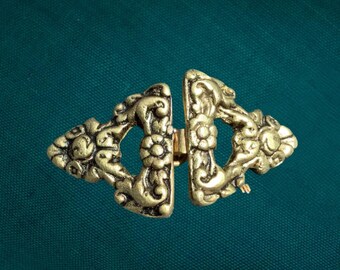Puede incluir: Un broche para capa de metal en tono latón compuesto por dos piezas triangulares simétricas. El cierre presenta detallados patrones florales y de volutas en relieve, junto con un mecanismo de bisagra central metálico.