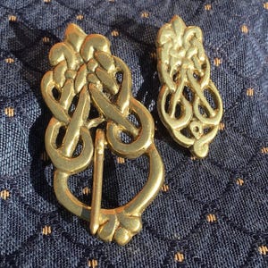 Puede incluir: Una hebilla de metal dorado y una pieza decorativa más pequeña a juego, ambas con un intrincado diseño de nudos entrelazados. La hebilla incluye un perno central y un marco redondeado, mientras que la pieza más pequeña tiene un patrón ornamental similar.