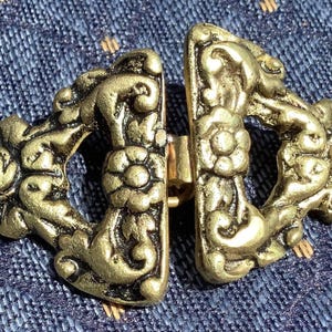 May include: A gold-toned metal clasp with intricate floral designs. The clasp is made of two pieces that fit together.