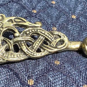 May include: A gold-toned metal belt buckle featuring an intricate design of a dragon or serpent intertwined with a knot. The buckle is detailed with raised lines and a smooth finish.