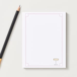 May include: A white notepad with a pink border and a small cocktail illustration in the corner. A black pencil with a gold ferrule rests on the left side of the notepad, creating a simple, elegant composition.