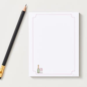 May include: A white notepad with a pink border and a champagne bottle and glasses illustration. A black pencil with a gold ferrule and eraser is positioned to the left of the notepad.