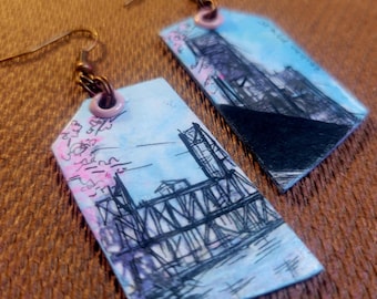 May include: Handmade tag-shaped earrings featuring a bridge design. The earrings have a light blue background with a black and purple bridge illustration, pink floral accents, and bronze-colored ear wires.