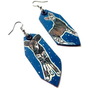 May include: A pair of blue and white dangle earrings featuring a painted image of an okapi, a type of African mammal, on each earring.