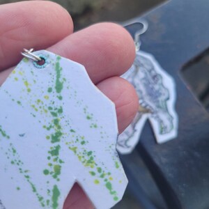 May include: A pair of earrings with a white background and green and yellow paint splatters. One earring is shaped like a dinosaur head, the other is a more abstract shape.