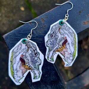 May include: A pair of silver earrings with white, green, and yellow dinosaur skull designs. The earrings are made of metal and have a hook closure.