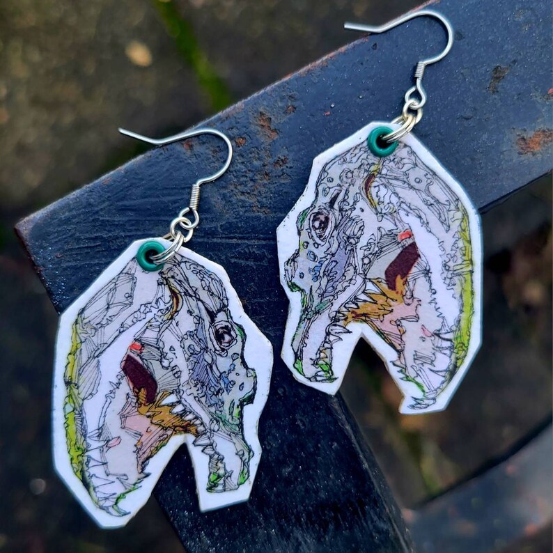 May include: A pair of silver earrings with white, green, and yellow dinosaur skull designs. The earrings are made of metal and have a hook closure.