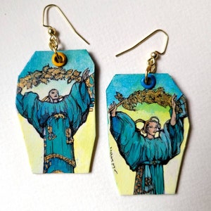 May include: A pair of earrings with a yellow and blue painted design featuring a figure holding a branch of leaves. The earrings are made of wood and have gold metal hooks.