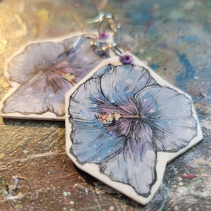 May include: A pair of earrings featuring a watercolour-style hibiscus flower design. The flowers are painted in shades of blue, purple, and white, with a hint of pink in the centre. The earrings are made of white ceramic and have silver hooks.