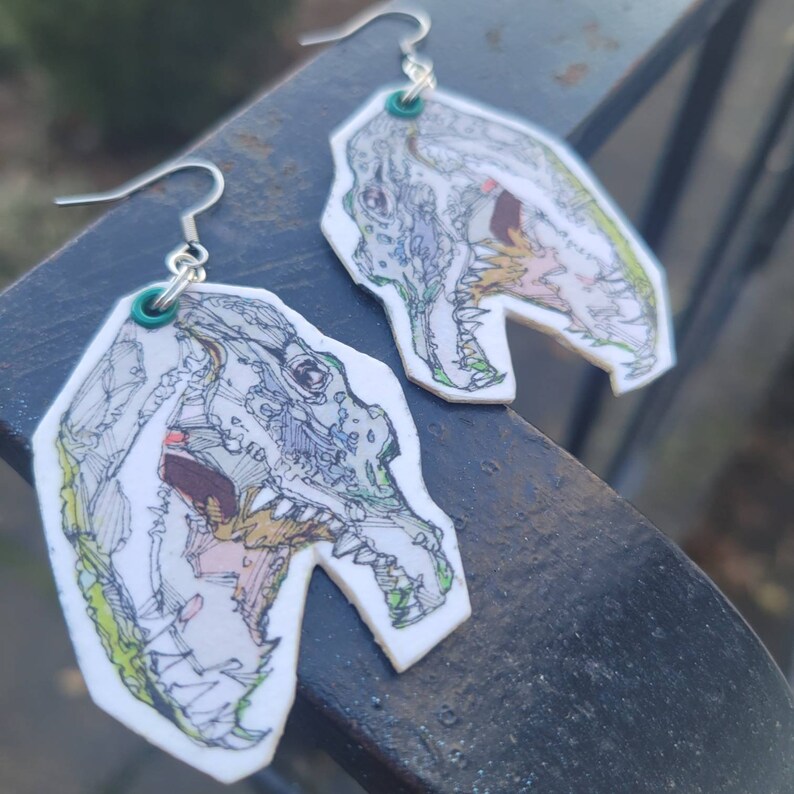 May include: A pair of silver earrings with a white background and a detailed black and white drawing of a dinosaur head. The dinosaur's mouth is open and its teeth are visible. The earrings are hanging from a silver hook.