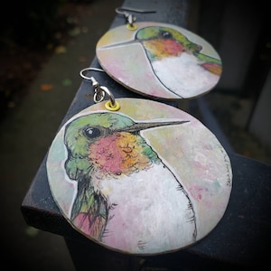 May include: A pair of round wooden earrings with a watercolor painting of a hummingbird on each. The hummingbird is green, pink, and white with a long beak. The earrings have silver hooks and yellow accents.