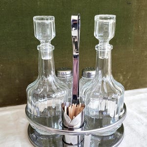 May include: A vintage glass condiment set with two bottles, two shakers, and a toothpick holder. The set is held in a silver-toned metal stand. The bottles have stoppers and the set sits on a round tray.