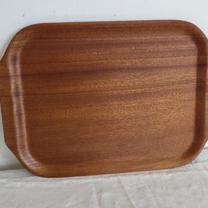 Puede incluir: Una bandeja de madera rectangular con esquinas redondeadas y bordes elevados. La bandeja está hecha de madera de color marrón rojizo con un patrón de vetas visible. La bandeja está vacía y se encuentra sobre una superficie blanca.