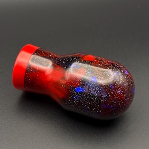 May include: A red and black bottle stopper with a galaxy-like design. The stopper has a red top and a black base with blue and silver glitter embedded in the resin.
