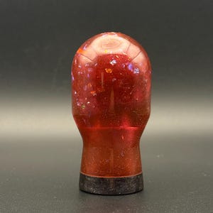 May include: A red and orange glass bottle stopper with a black base. The glass is filled with iridescent glitter.
