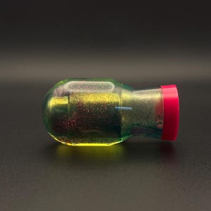 May include: A clear, yellow, green, and pink bottle with a pink lid. The bottle is filled with a glittery liquid.