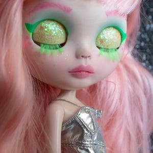 Pode incluir: Uma boneca com cabelo rosa e um vestido prateado. A boneca tem olhos grandes com íris amarelas brilhantes, delineador verde e cílios verdes. Os lábios da boneca são de um rosa suave.