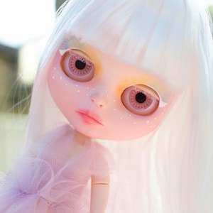 Pode incluir: Um close-up de uma boneca com tez pálida, olhos rosa grandes e cabelo branco. A boneca tem um vestido rosa e pequenas sardas brancas no rosto. Os olhos da boneca têm uma íris detalhada e uma pupila preta.